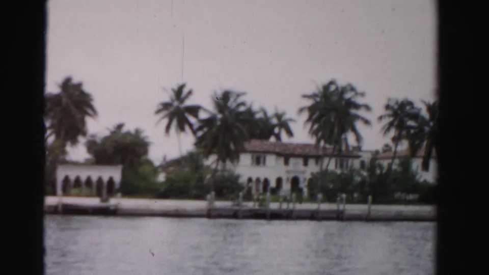 Soft 8mm frame of a white Dade County waterfront house with palm trees, landscaping, dock posts, and water in front.