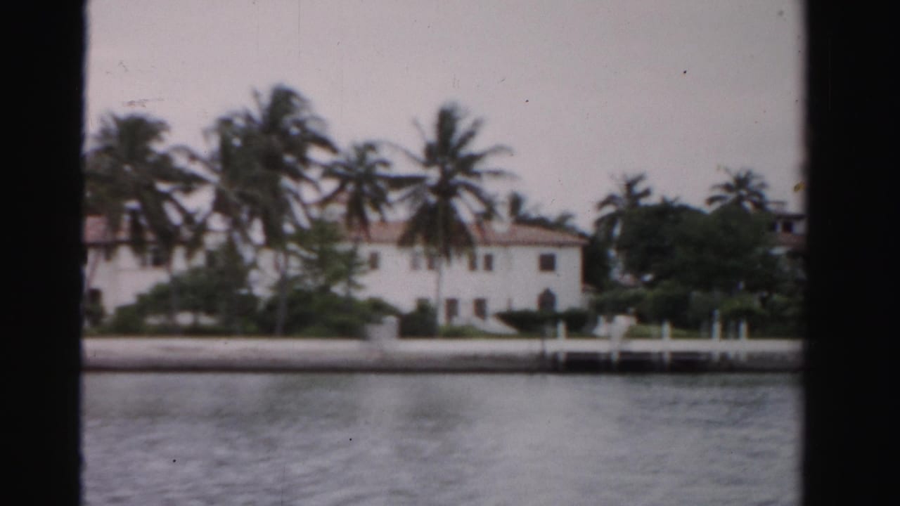1955 Dade County 8mm frame showing a white waterfront house, palms, a seawall, and calm water across the foreground.