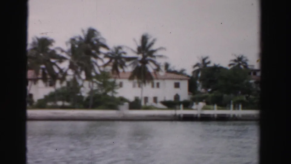 1955 Dade County 8mm frame showing a white waterfront house, palms, a seawall, and calm water across the foreground.