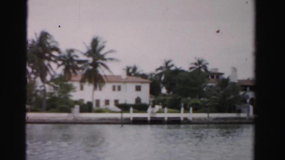 1955 Dade County view of waterfront houses framed by palms, white dock railings, and rippled water in soft 8mm color.