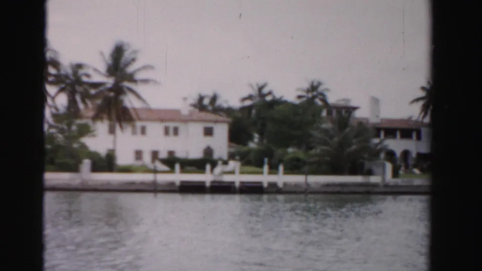 A 1955 Dade County frame centers on a white waterfront residence with palms, dock posts, seawall, and water in the foreground.