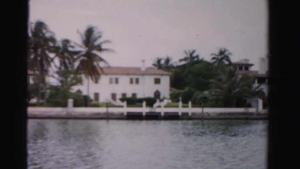1955 Dade County home-movie frame with a white waterfront home, palms, dock, seawall, and calm water under faded color.