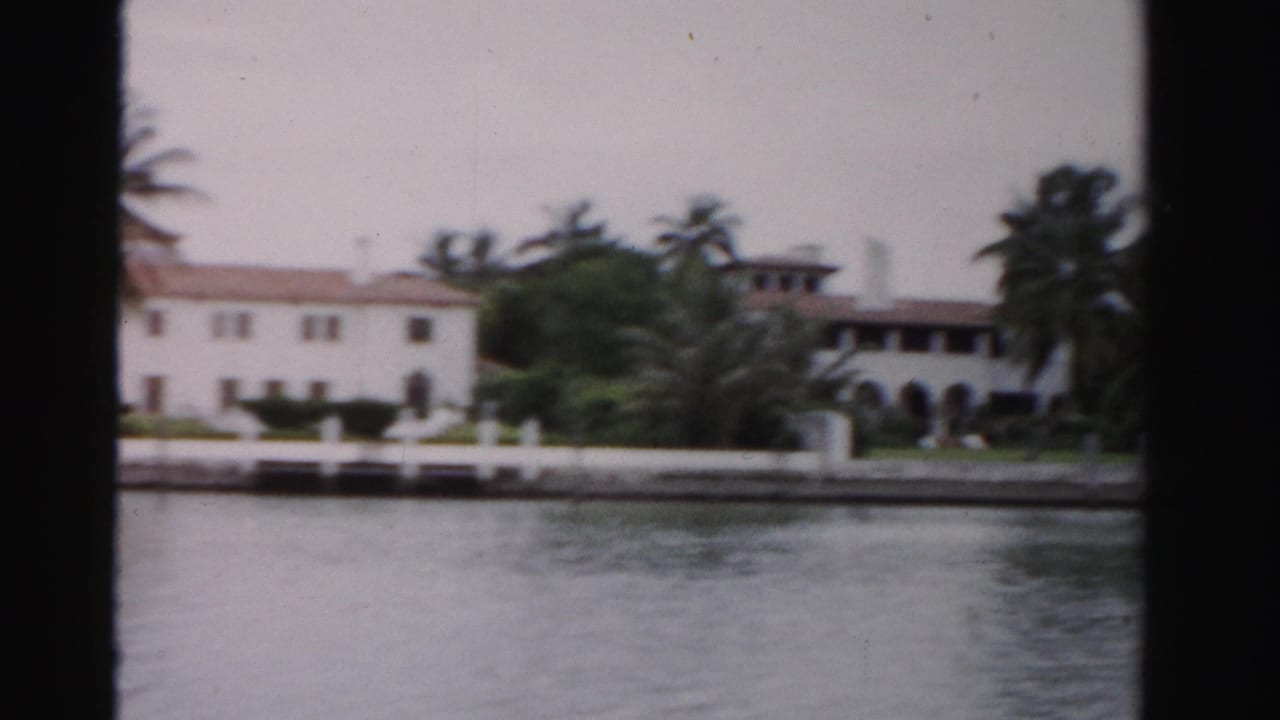 1955 Dade County 8mm view of a white waterfront building with red tile roof, palms, lawn, and seawall seen from across the water.
