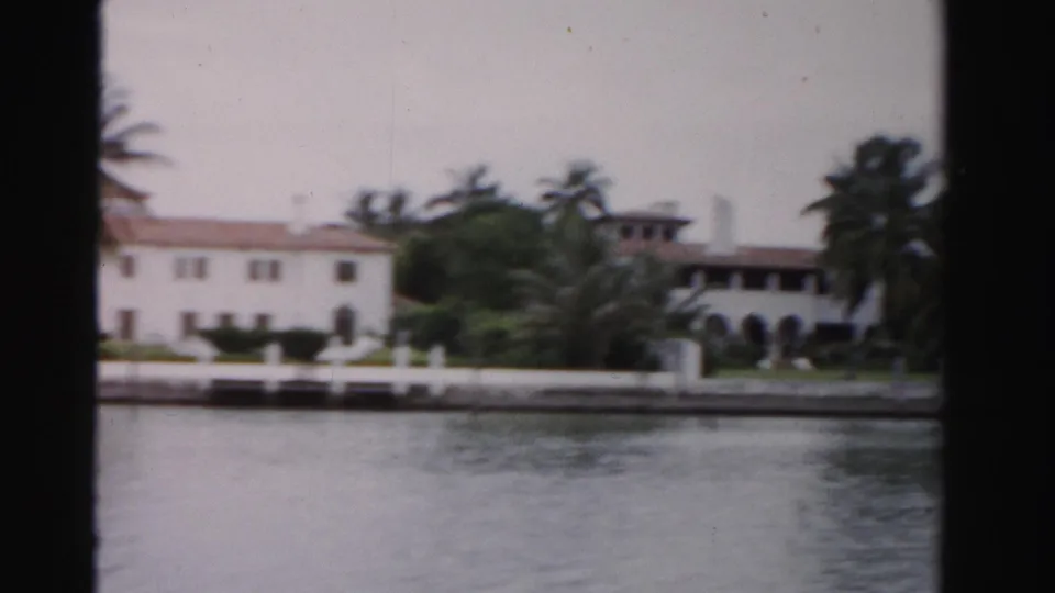 1955 Dade County 8mm view of a white waterfront building with red tile roof, palms, lawn, and seawall seen from across the water.