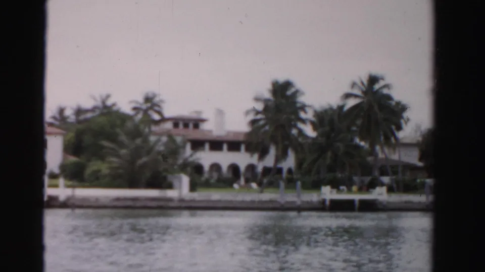 1955 Dade County waterfront view showing a white Spanish-style structure, palms, arched loggia, seawall, and small dock area.