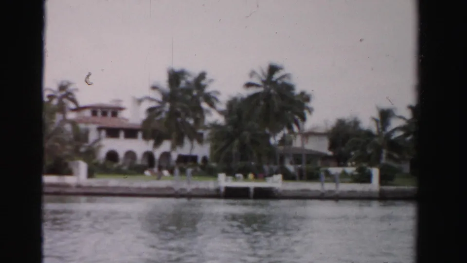 1955 Dade County 8mm frame of a waterfront Spanish-style building with palms, arched openings, lawn, seawall, and dock details.