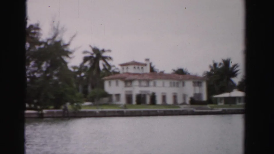 1955 Dade County view of a white waterfront house or tourist structure with red tile roof, palms, manicured lawn, and seawall.