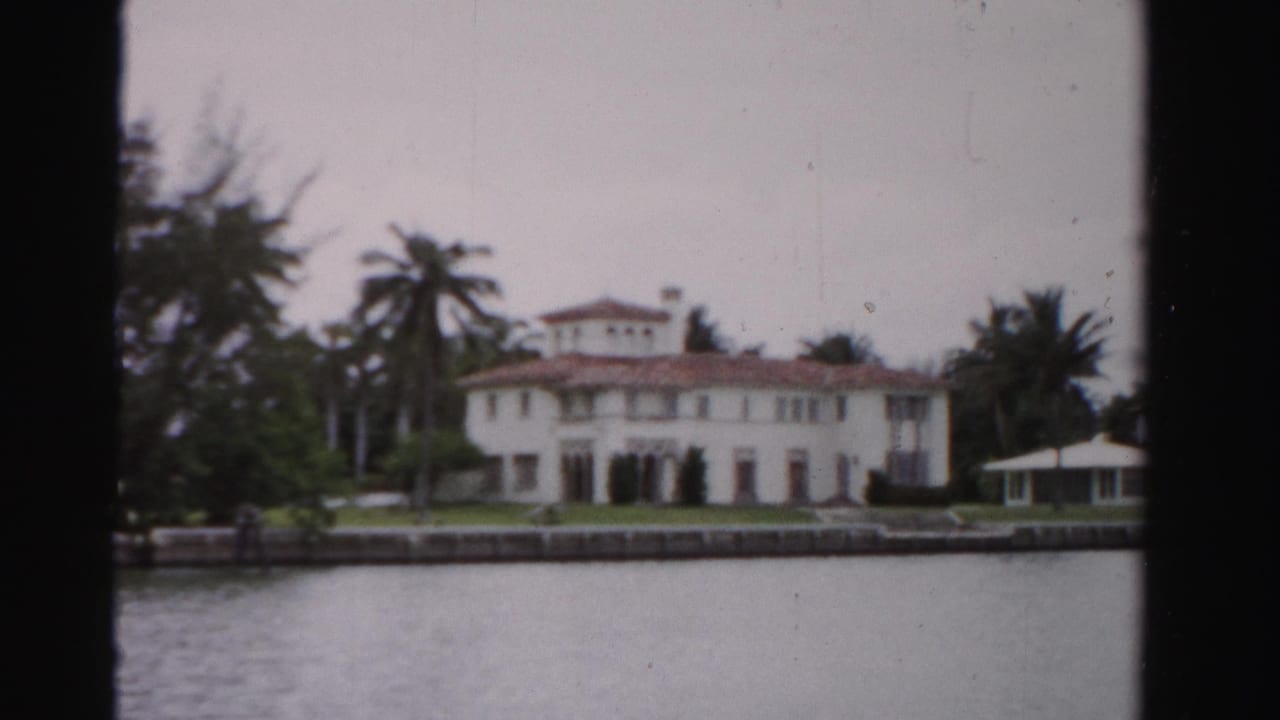 White waterfront mansion with palms, seawall, lawn, and small gazebo across calm water.