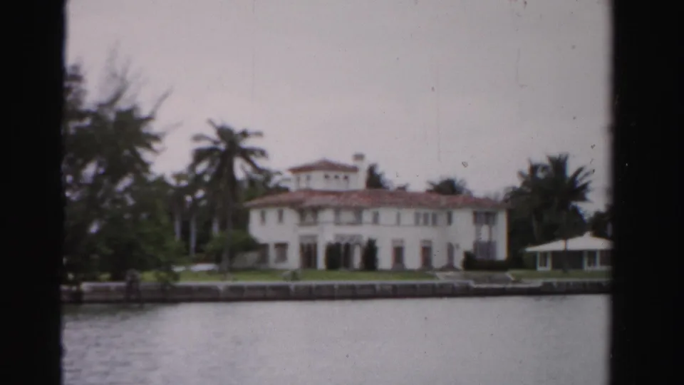 1955 Dade County, Florida home-movie view of a white Mediterranean-style waterfront mansion with palms, seawall, lawn, and gazebo.