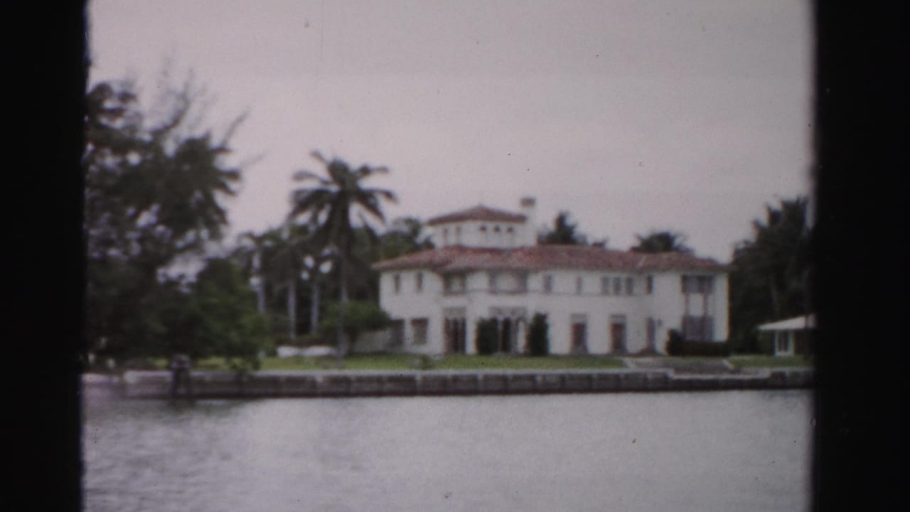 1955 Dade County, Florida view of a white waterfront estate with terracotta roof, palm trees, seawall, lawn, and small gazebo.