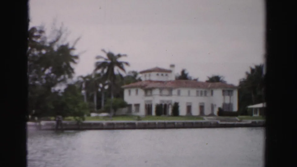 1955 Dade County, Florida view of a white waterfront estate with terracotta roof, palm trees, seawall, lawn, and small gazebo.