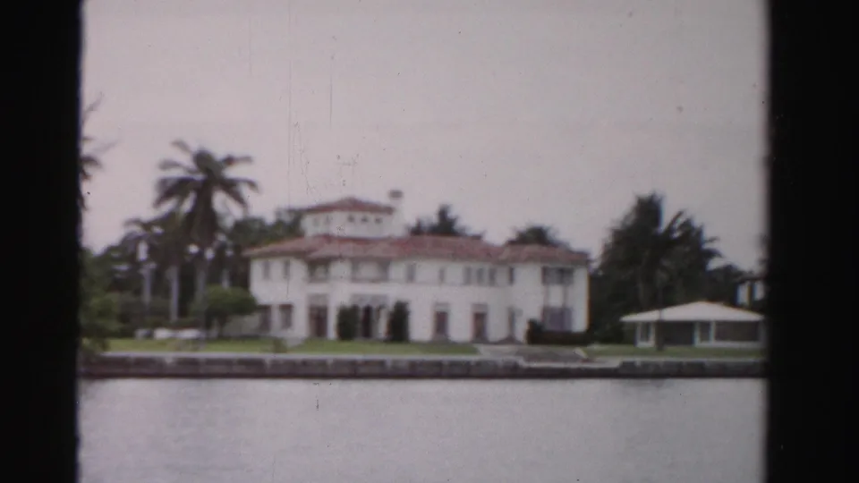 Soft 8mm frame from 1955 Dade County showing the waterfront mansion, palms, green lawn, seawall, and gazebo structure.