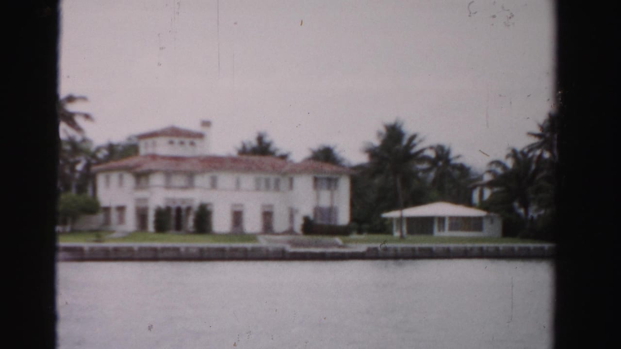 White waterfront mansion with red-tile roof, palm trees, lawn, seawall, and calm water.