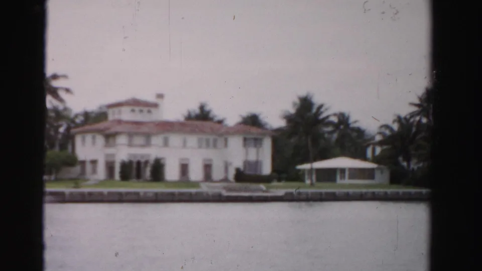 1955 Dade County waterfront estate seen from across the water, with a white Mediterranean-style house, palms, lawn, and seawall.