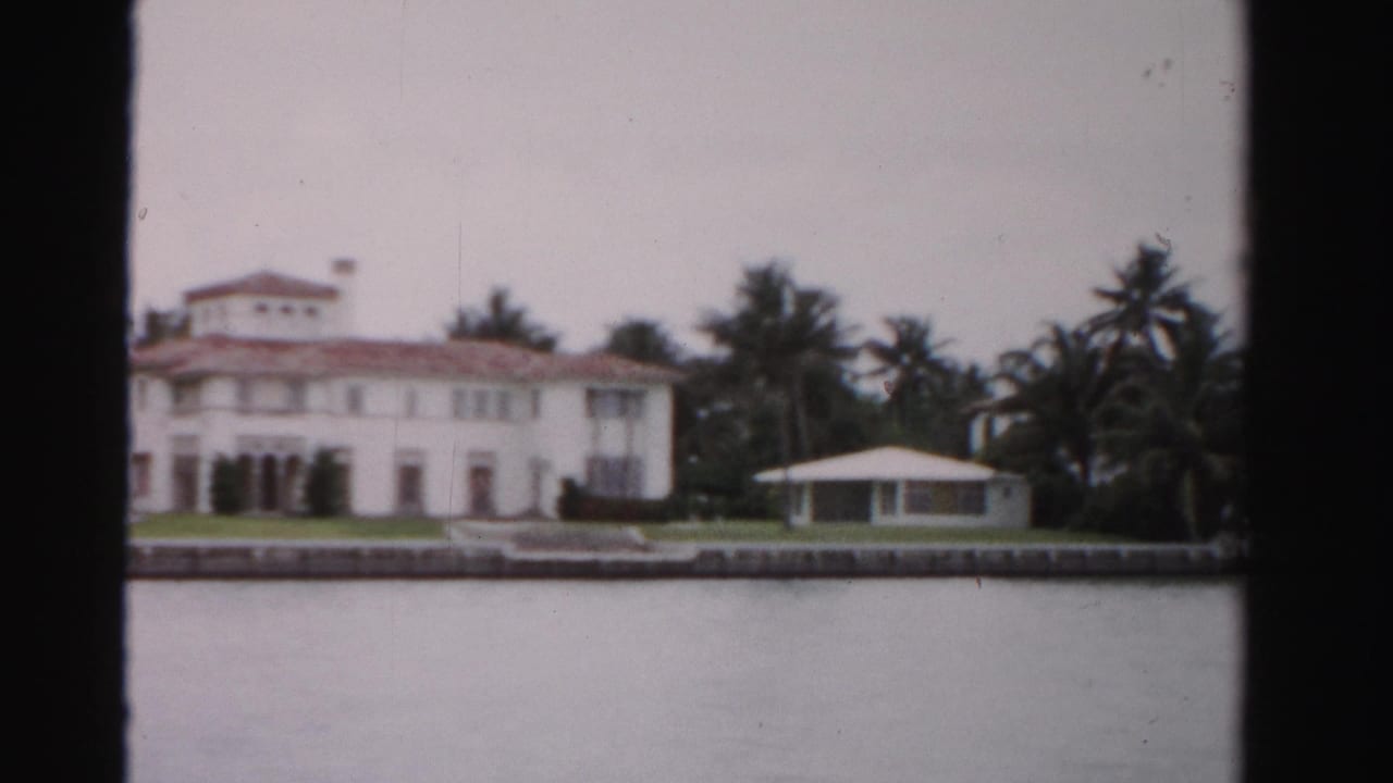 1955 Dade County estate view showing the main white house, red roof, palms, low seawall, and a small pavilion-like guest house near the water.