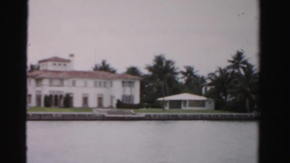 1955 Dade County estate view showing the main white house, red roof, palms, low seawall, and a small pavilion-like guest house near the water.