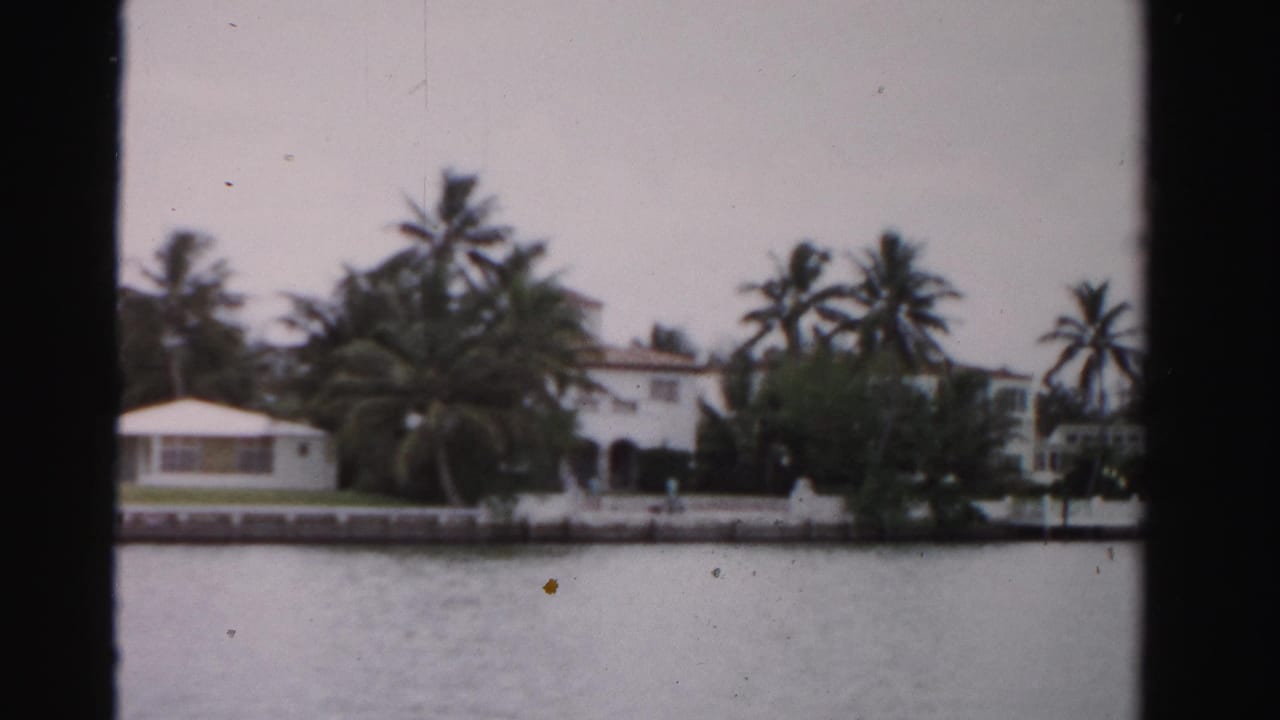 1955 Dade County waterfront estate view with white stucco buildings, palms, seawall, and calm water seen from across the shore.