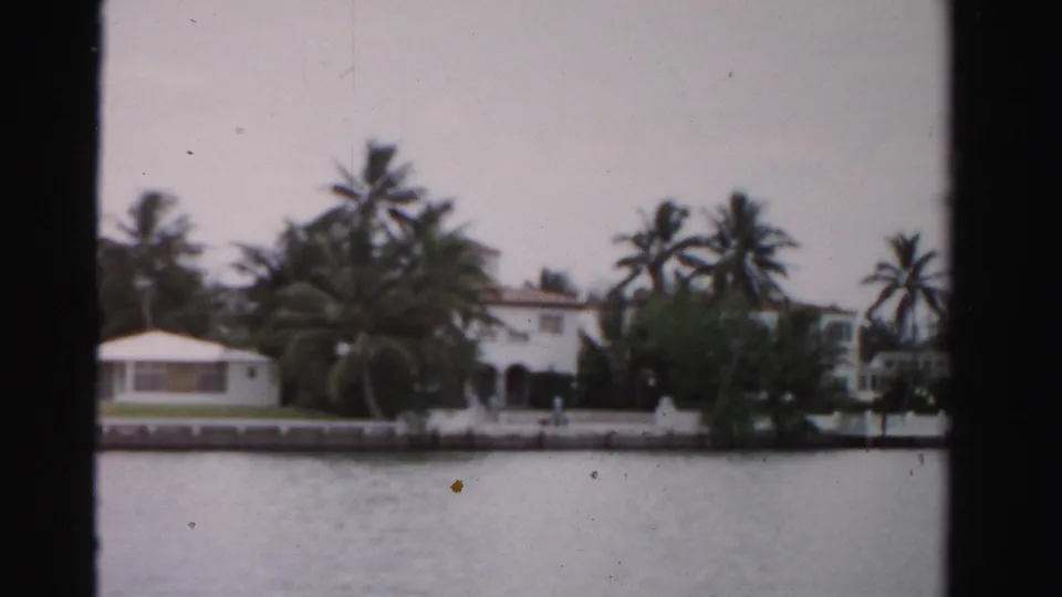 1955 Dade County waterfront estate view with white stucco buildings, palms, seawall, and calm water seen from across the shore.