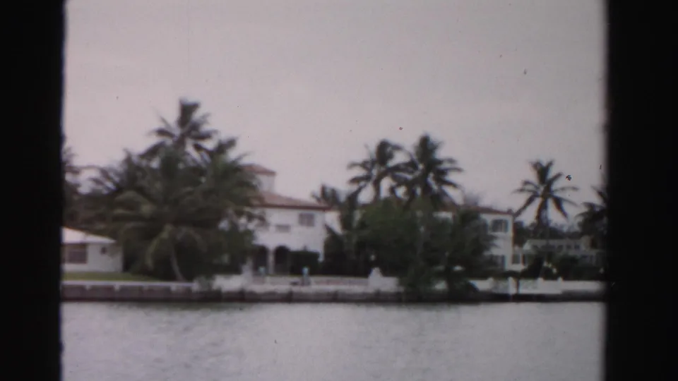 1955 Dade County shoreline view of a large white residence partly screened by palms and greenery, with water in the foreground.