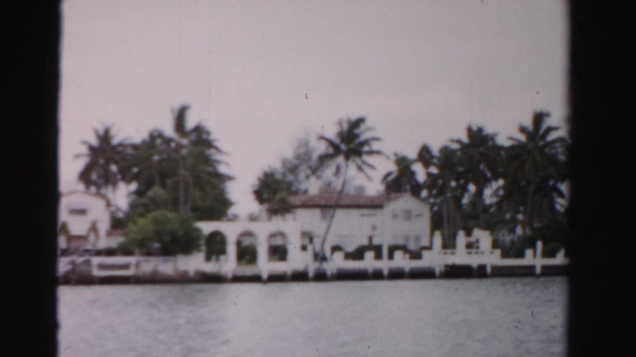 1955 Dade County waterfront view of a white Mediterranean-style house, palms, seawall, and rippled water.