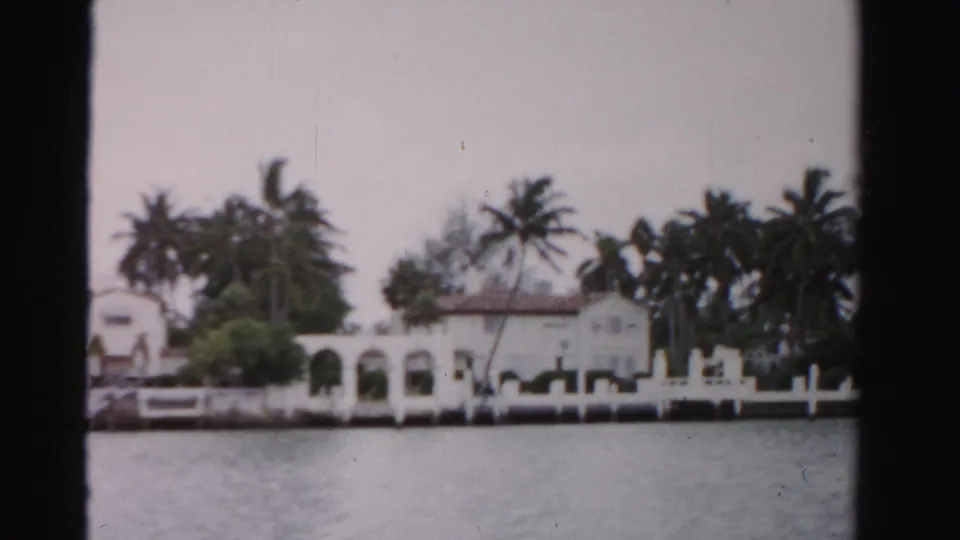 1955 Dade County waterfront view of a white Mediterranean-style house, palms, seawall, and rippled water.