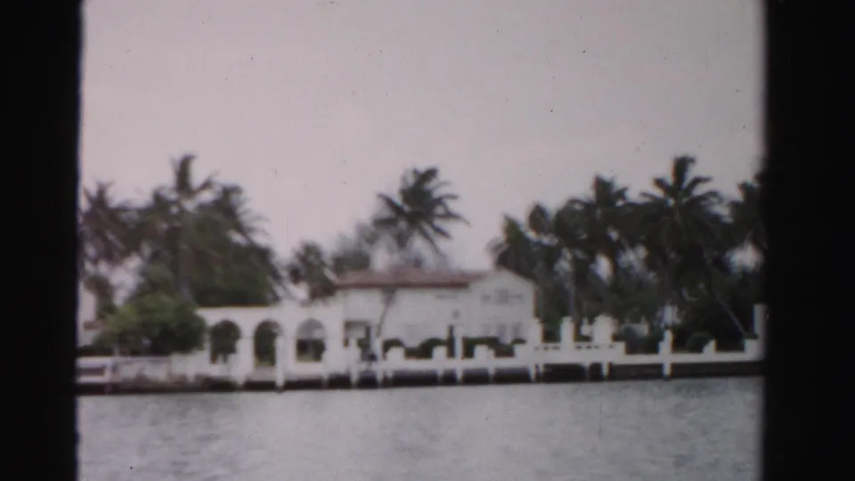 Moving 8mm view toward a Dade County waterfront residence with palms, arched facade, seawall, and open water.