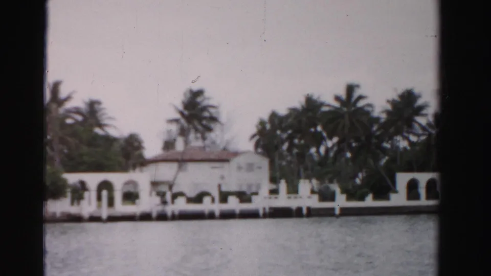 1955 Florida home-movie frame showing a white arched waterfront building, palms, seawall, and water in the foreground.