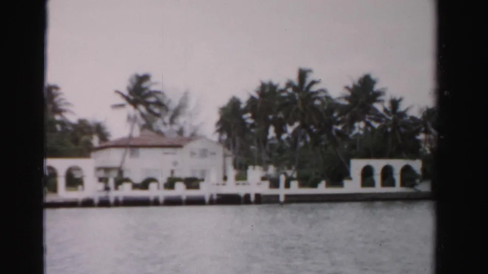 Dade County shoreline view with a white waterfront home, dense palms, arched side structure, seawall, and rippled water.