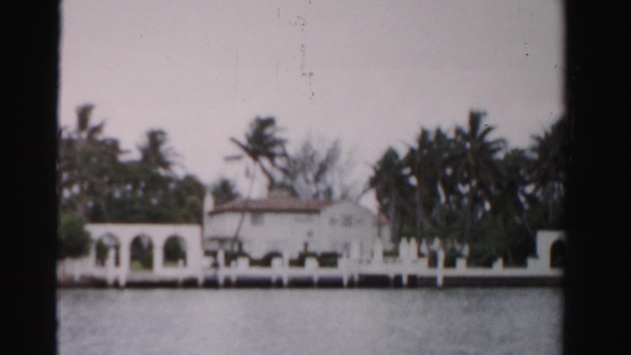 White waterfront house with arched openings and palm trees seen across water.