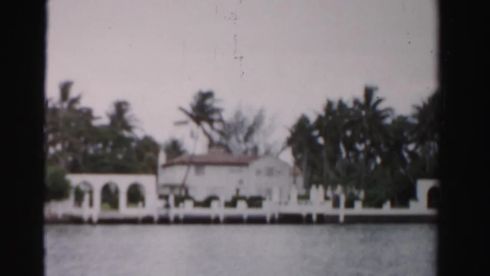 1955 Dade County 8mm frame showing a white Mediterranean-style waterfront house with palms and arched walkways.
