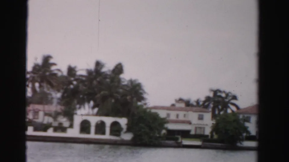 1955 Dade County 8mm frame showing white waterfront houses, arched openings, palms, and a low seawall beside calm water.