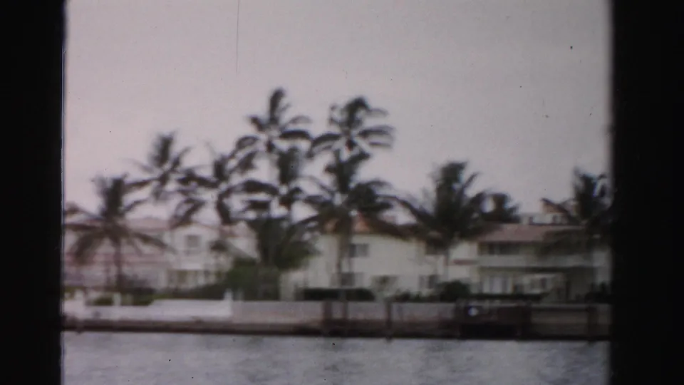1955 Dade County waterfront view with palms, white houses, a seawall, and a small dock seen from a moving camera.