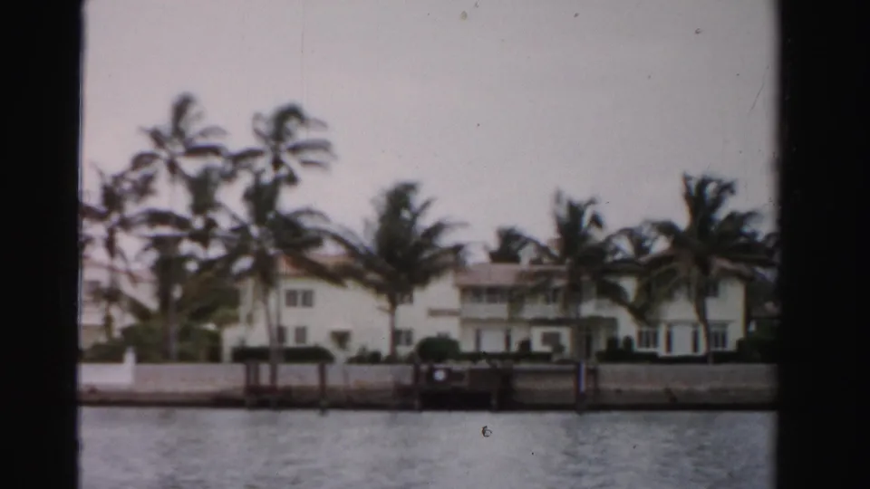 Dade County home-movie frame from 1955 showing a two-story waterfront residence, palms, seawall, and dock structure.