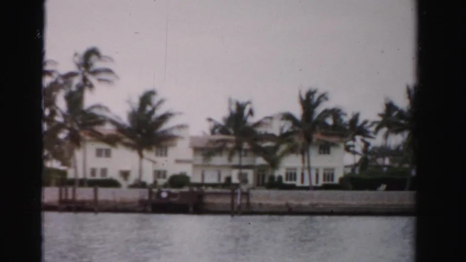 1955 Dade County 8mm frame with waterfront homes, palms, low seawall, dock structure, and rippled water in the foreground.