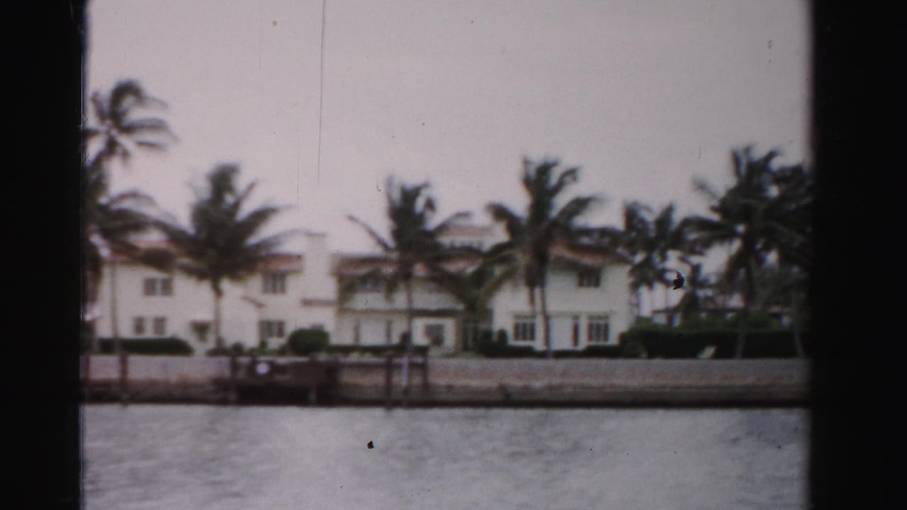 1955 Dade County 8mm view of white waterfront homes, palms, and a seawall seen from a boat on calm water.