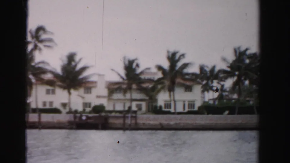 1955 Dade County 8mm view of white waterfront homes, palms, and a seawall seen from a boat on calm water.