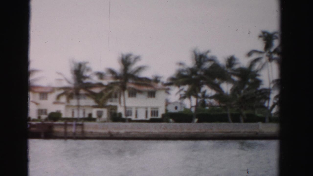 Long white waterfront house with palms and a seawall viewed across water.