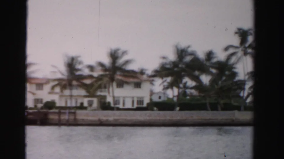 Boat-level 1955 Dade County view of a long white waterfront residence, palm trees, hedges, and a concrete seawall.