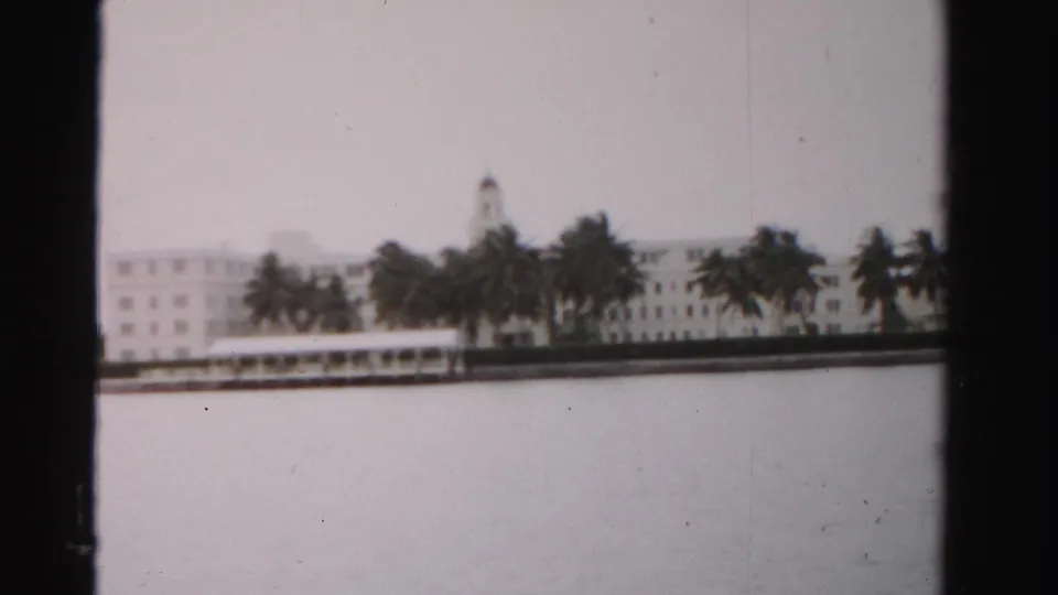 Faded 1955 Dade County 8mm view of white waterfront buildings partly screened by palms across pale water.