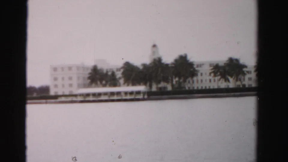 1955 Dade County home-movie frame showing a white waterfront building and palm trees seen across calm water.
