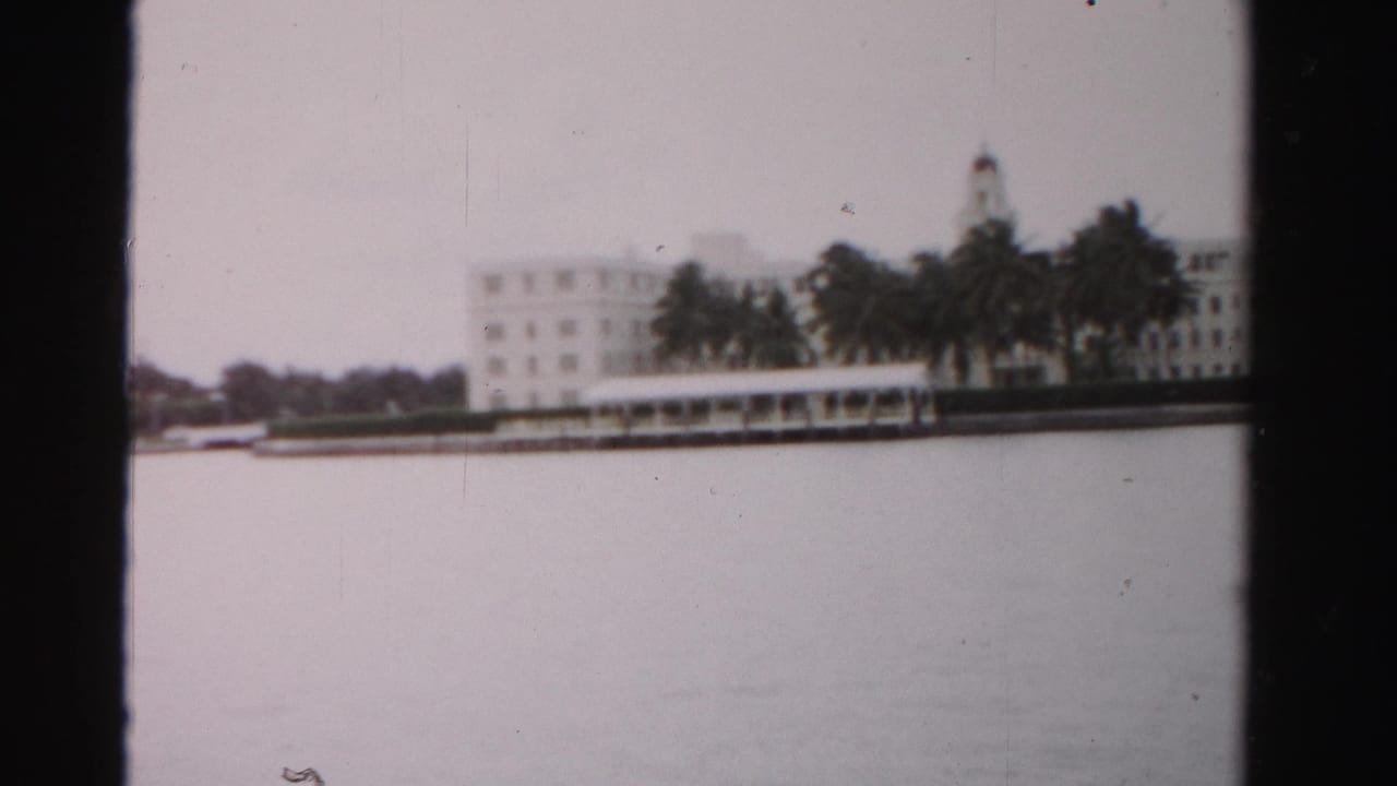 A 1955 Dade County waterfront view with a large pale building, palms, and still water in soft 8mm focus.