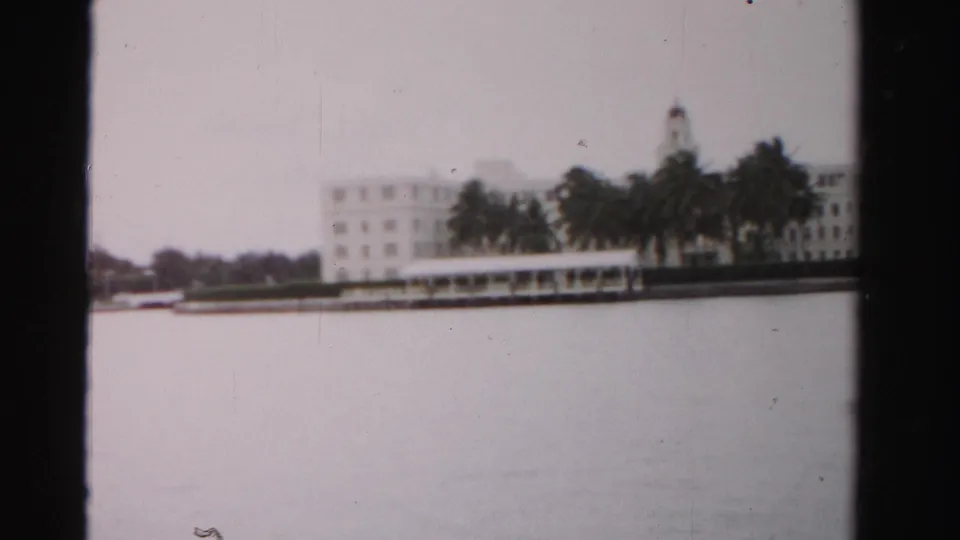 A 1955 Dade County waterfront view with a large pale building, palms, and still water in soft 8mm focus.
