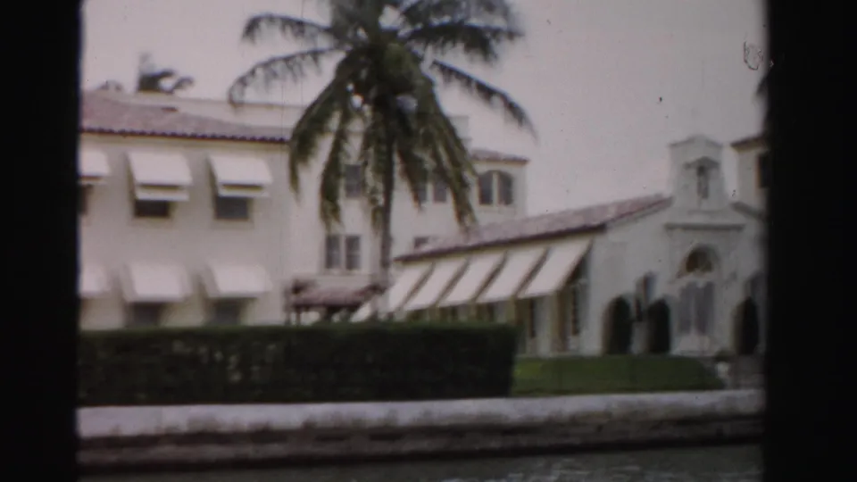 Closer 1955 Dade County frame showing a waterfront house facade with awnings, a large palm, hedge, and tower-like architectural detail.
