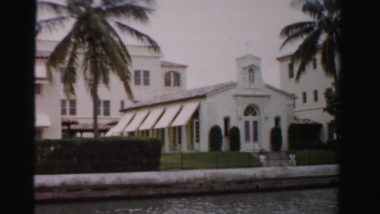 1955 Dade County home-movie frame showing a white canal-side church-like building, palms, lawn, hedges, and dark foreground framing.