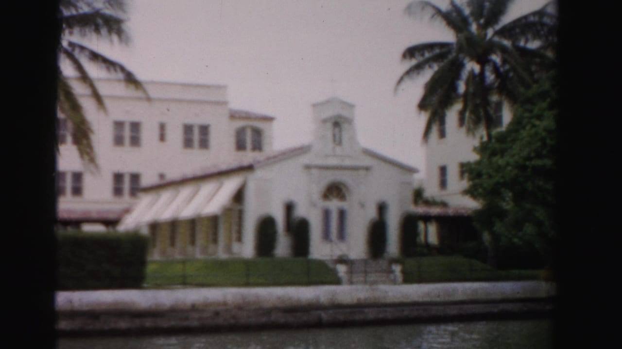 White waterfront church facade with awnings, palms, hedges, and canal edge in front.