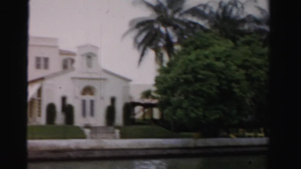 Soft 8mm frame from Dade County showing a partial white church-like building, palms, green shrubbery, lawn, steps, and canal foreground.