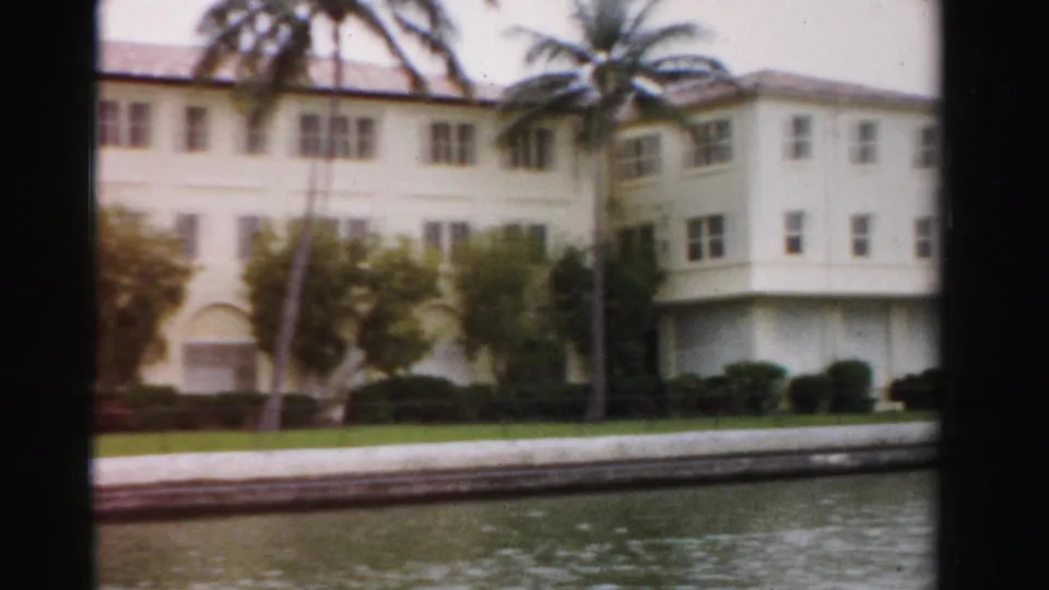 1955 Dade County 8mm frame showing the building facade closer, with palms crossing the view and lake water below.