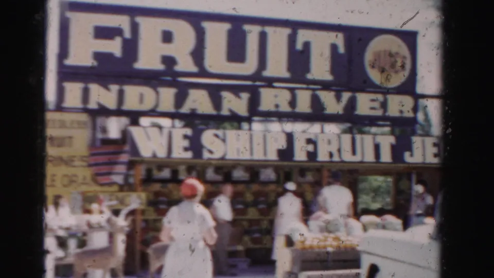 1955 Dade County frame of the Indian River Fruit Shop area, with large fruit and jelly signs, adult visitors, produce displays, deer, and vehicles.