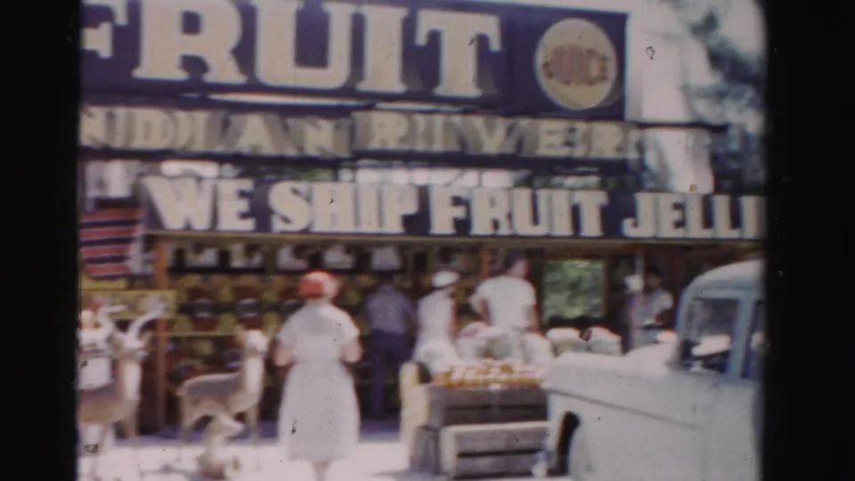 1955 Dade County home-movie frame showing the fruit shop frontage with bold signage, adults near the counter, produce, deer, and a parked vehicle.