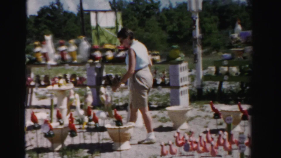 The woman leans toward small outdoor sale items, with red lawn figures, ceramic birds, and display tables visible in the 8mm frame.